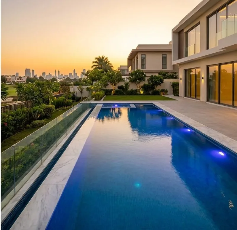 The same pool after renovation, with deep blue tiling, a white marble deck, a glass balustrade and underwater lighting at dusk
